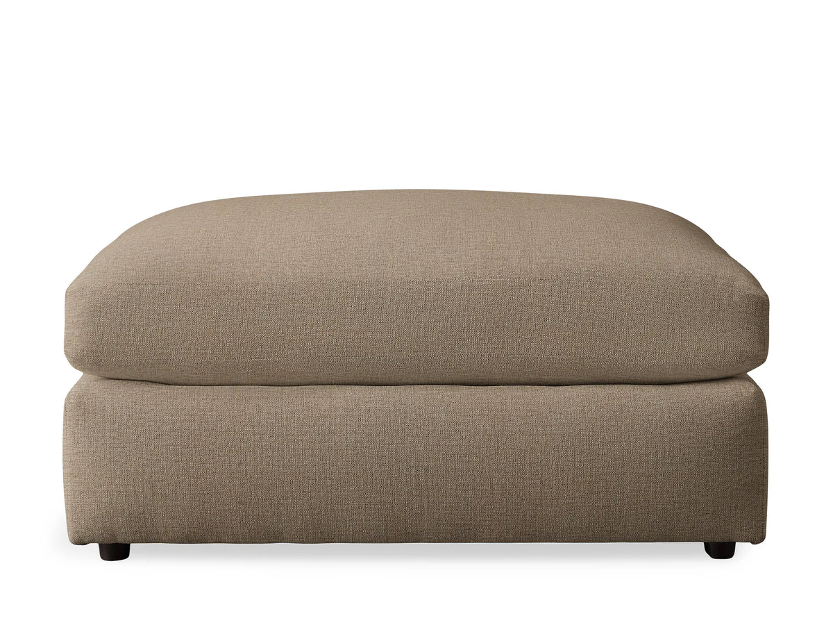 A Kipton Ottoman in soft, light brown textured fabric, featuring a plush, rounded top cushion and a sturdy rectangular base with small dark feet.