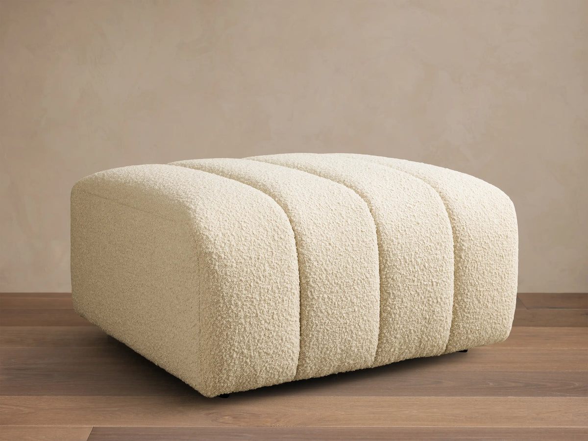 Front view of a cream Bracco Ottoman with a ribbed design and boucle texture, resting on a wooden floor.