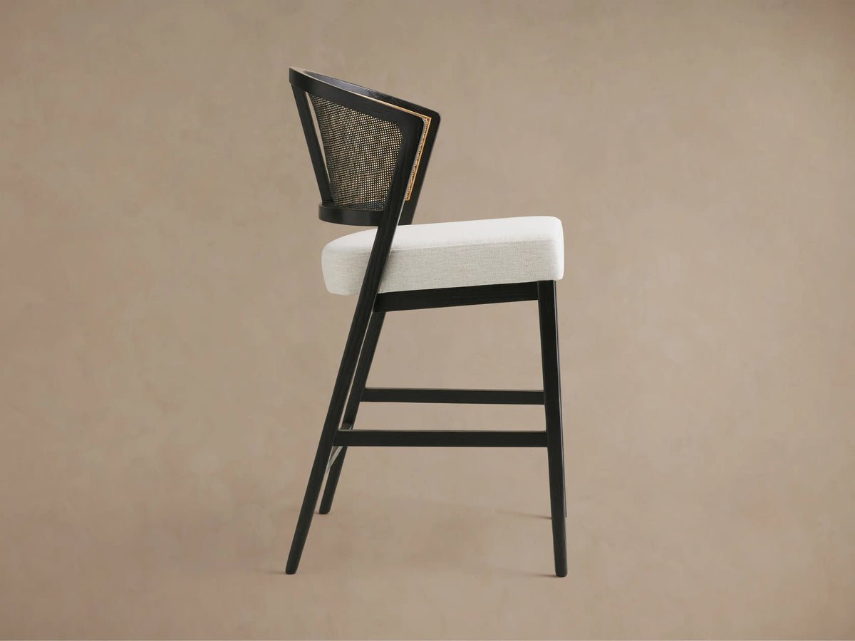 Aimee Armless Counter Stool with a solid oak frame, rattan cane back, and upholstered seat in a light fabric, shown from the back.