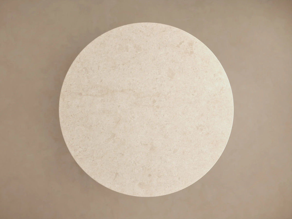 Close-up of Caden Round Dining Table. Round, light beige stone top with subtle texture, resting on a rich brown wood cylindrical base.