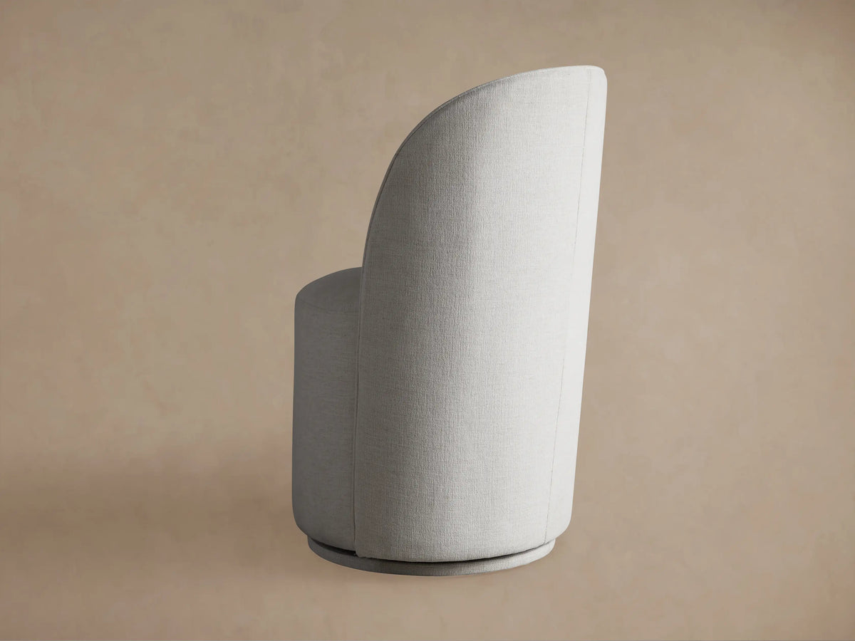 Kira Swivel Dining Chair in Nomad Snow fabric, side view, with curved back and cylindrical base, against a subtle brown wall.