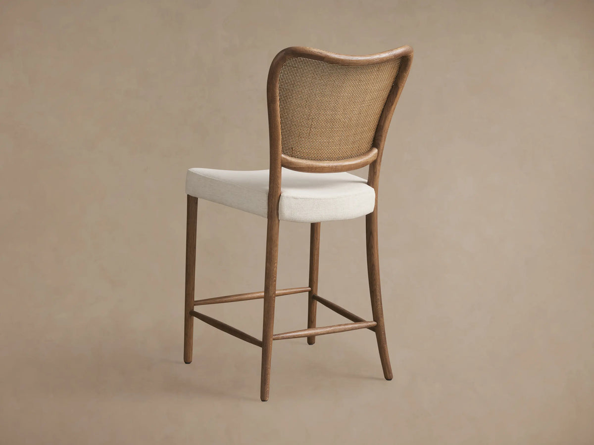 Close-up of Noa Stool in Cinder, showcasing its curved brown oak frame, woven rattan backrest, and textured off-white fabric seat.