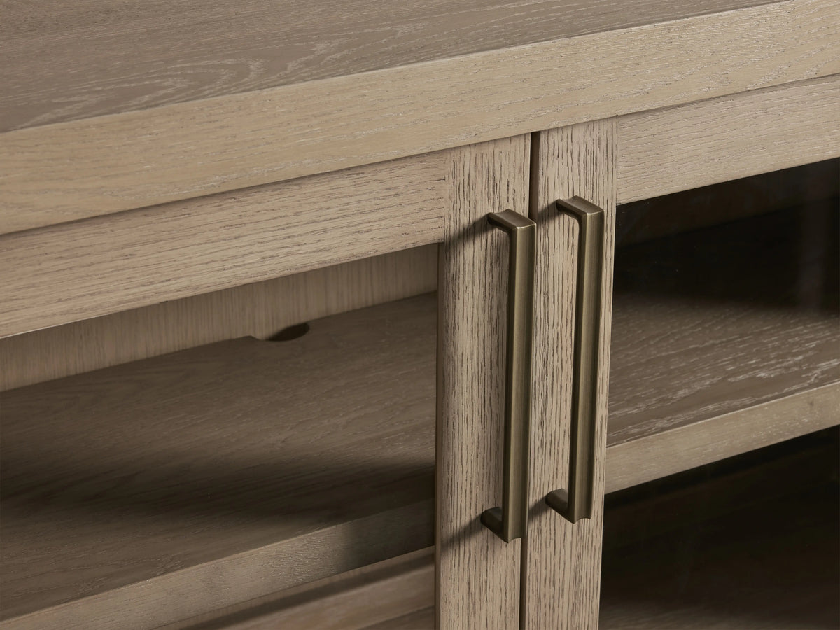 Close-up of Bodhi Media Console showing light oak wood grain, brass handles, and adjustable shelves behind glass doors.