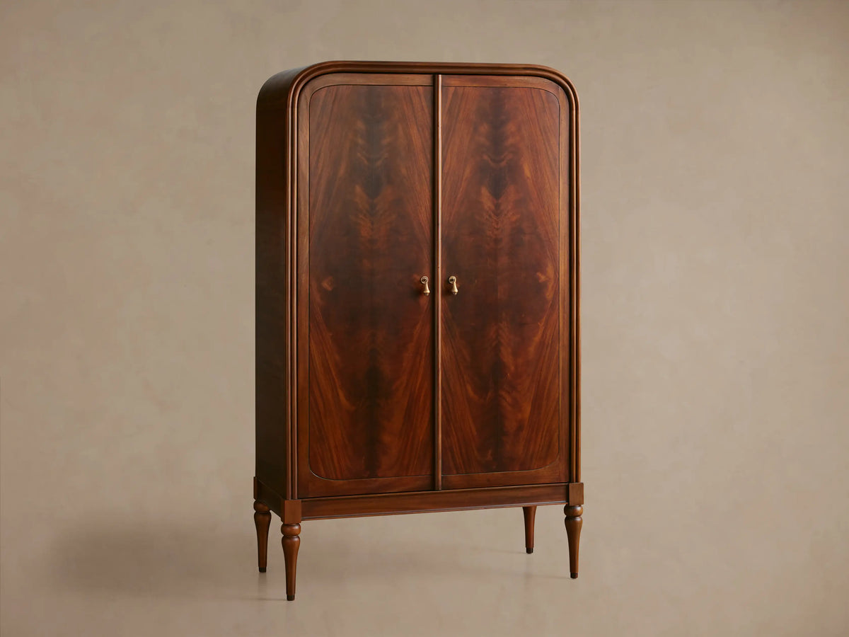 Close-up of Frances Cabinet's dark mahogany door with figured grain, brass handle, and interior shelves with books and bowl.