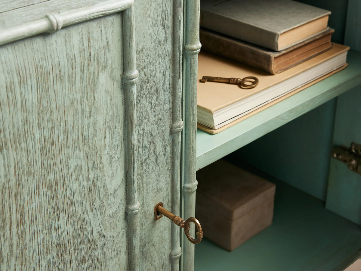 Iris Hall Chest close-up: Patinated teal-green finish, bamboo framing. Open drawer reveals books & a box, with a bronze pull.