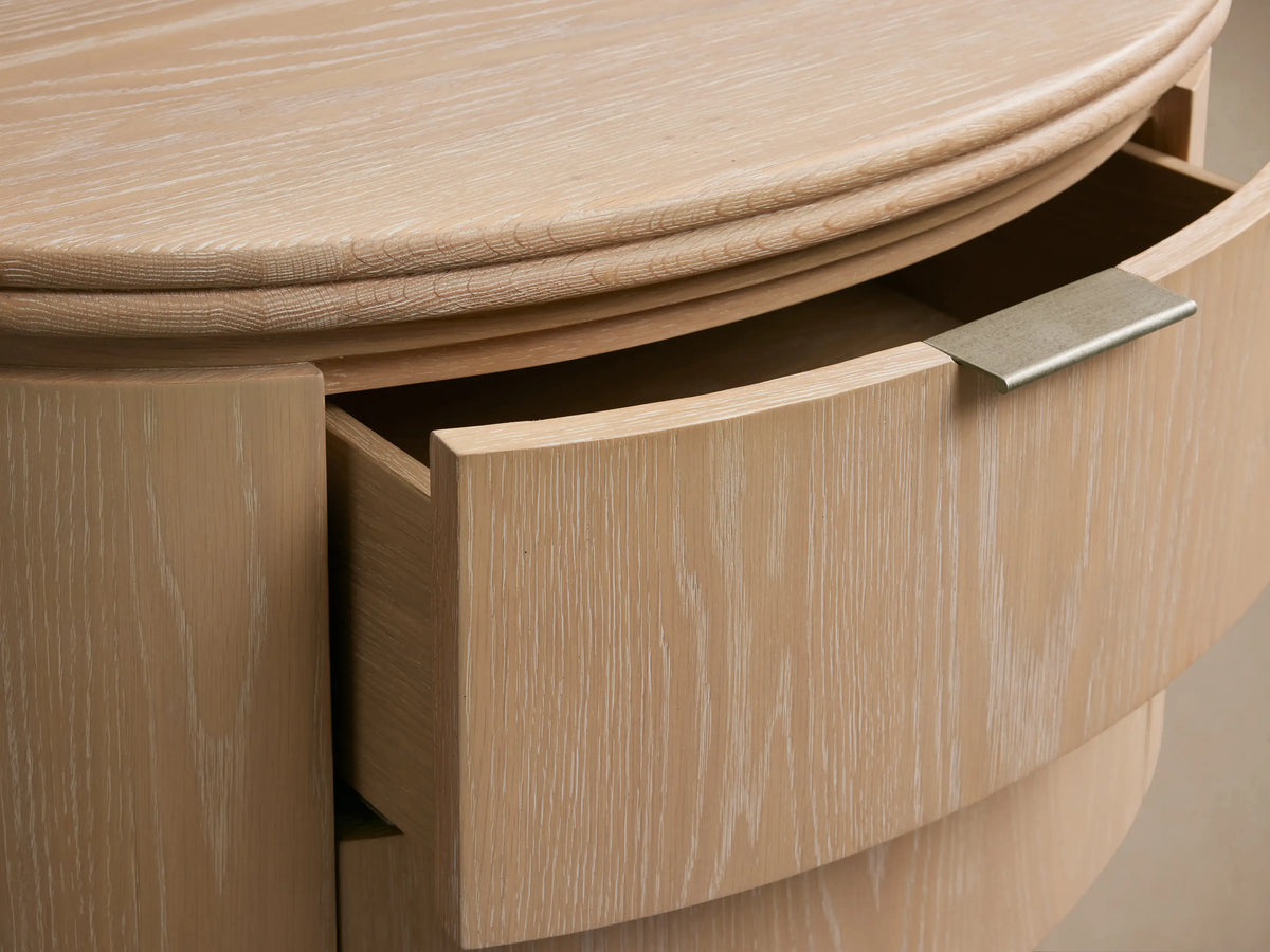 Caden Wood Oval Nightstand in sand finish oak veneer, shown with one curved drawer partially open, featuring pewter edge-pull hardware.