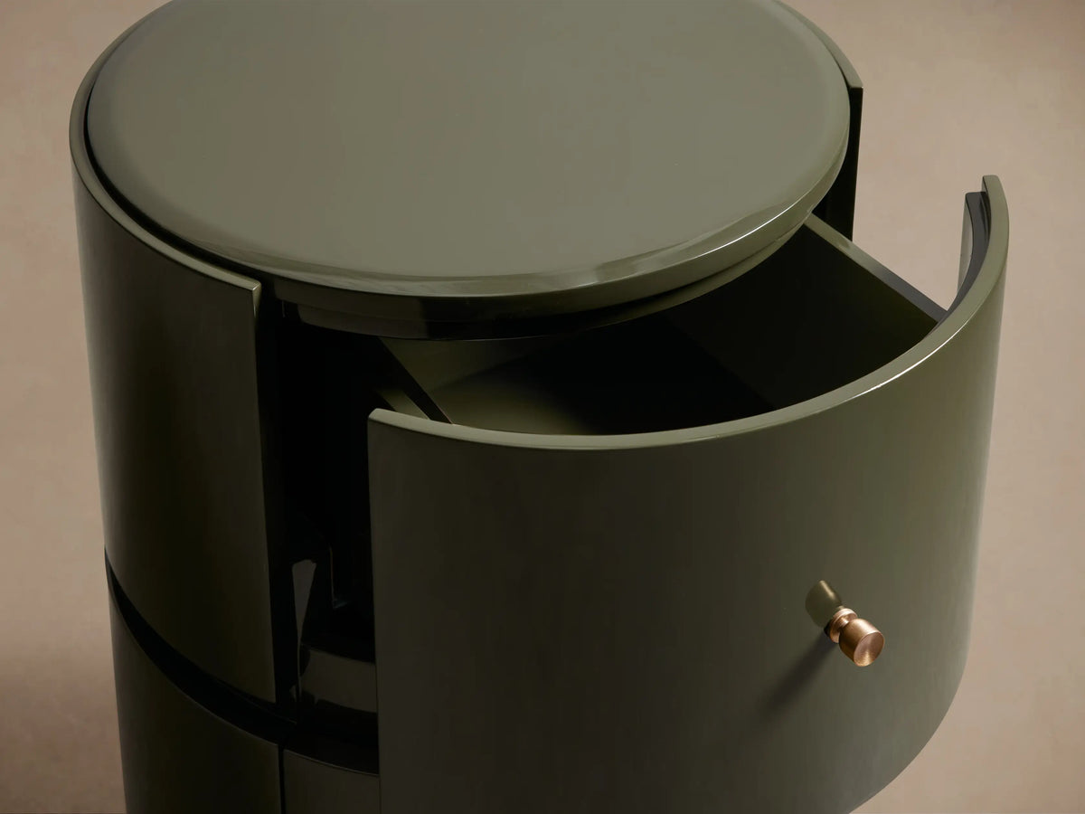 Dark green high-gloss Riya Round Nightstand with two stacked drawers and brass hardware, on a wood floor.