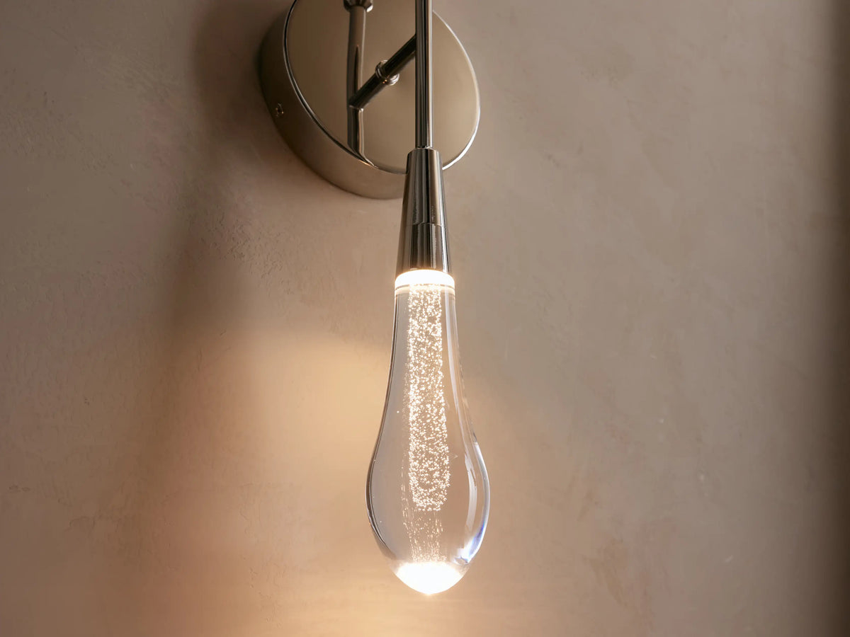 Soltaire Double Sconce with a nickel finish, featuring a teardrop-shaped glass bulb glowing warmly, mounted on a round backplate against a beige wall.