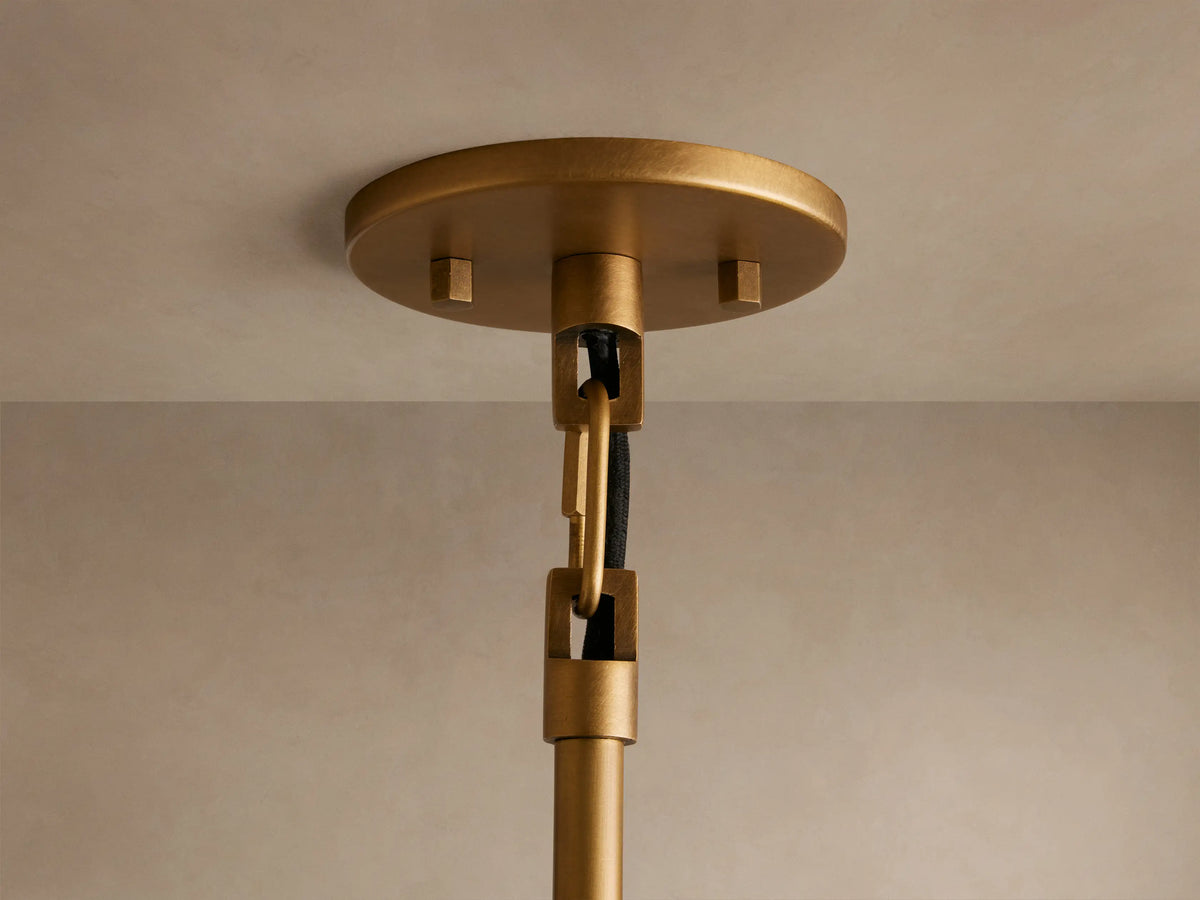 A sleek, brass pendant light with a wide, conical shade hangs vertically. Its exposed bulb casts a warm glow against a beige background.