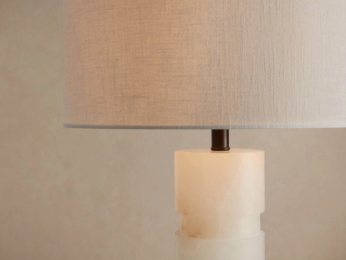 Novalie Table Lamp with a white cylindrical shade, carved alabaster base, and dark bronze bottom, on a wooden table.