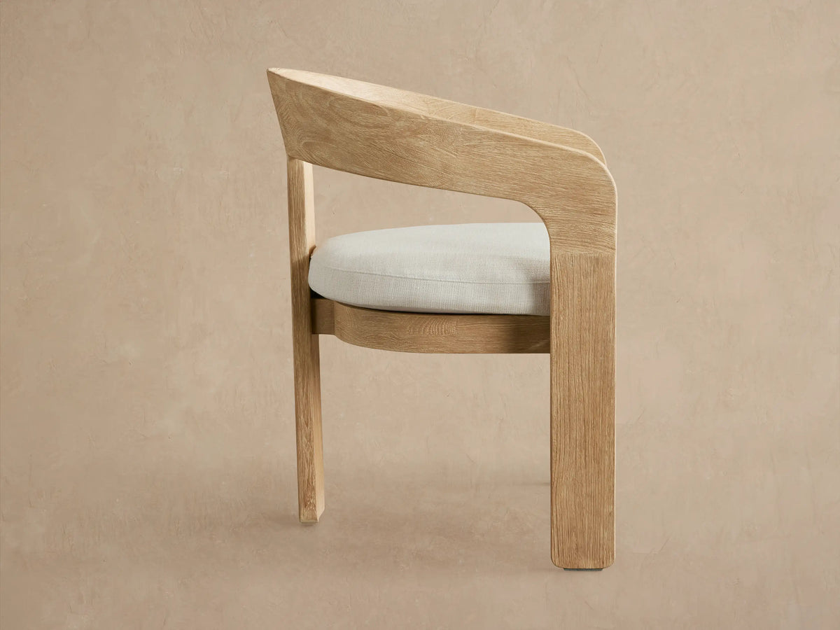 Anami Outdoor Dining Chair, light teak wood with curved back and arms, light beige seat cushion, angled view.