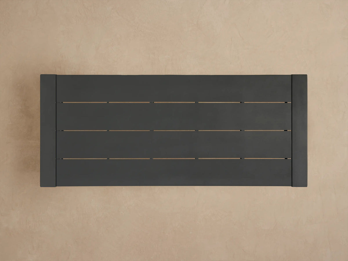 Dark gray Canyon Outdoor Aluminum Dining Table corner, showing a slatted top and thick square leg on a brown background.