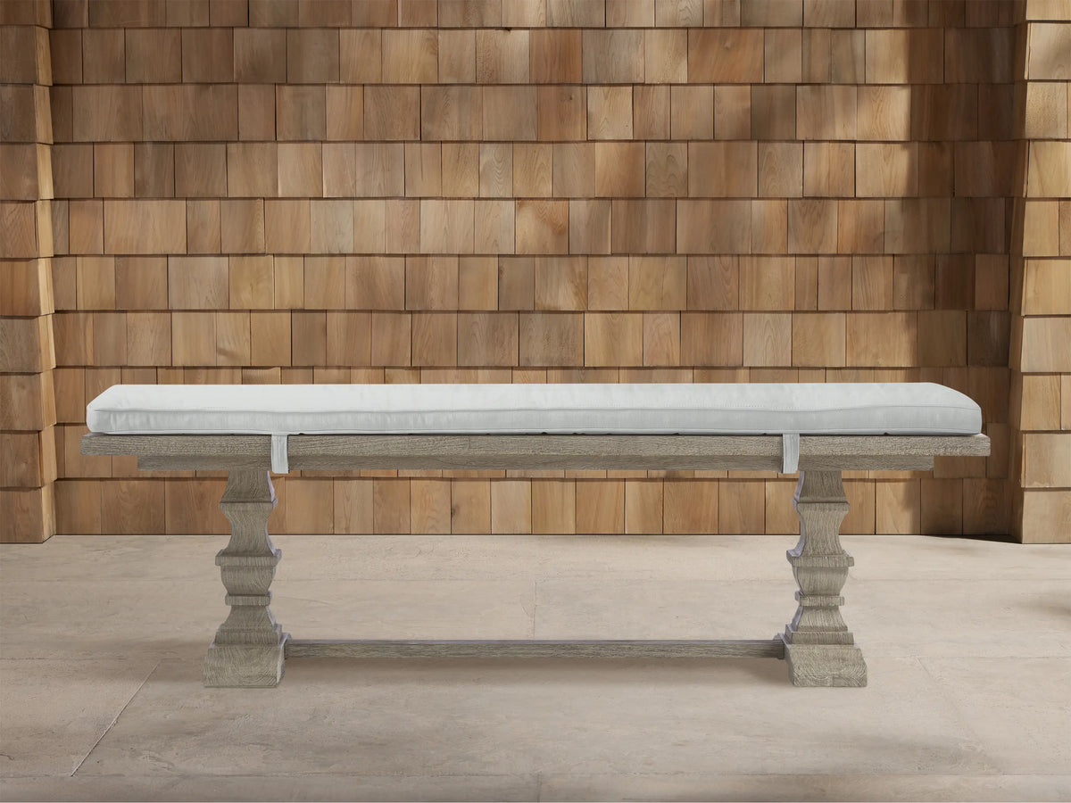 Weathered fawn Hamptons outdoor dining bench, carved pedestals, with a strapped light grey cushion, against a wooden shingle wall.