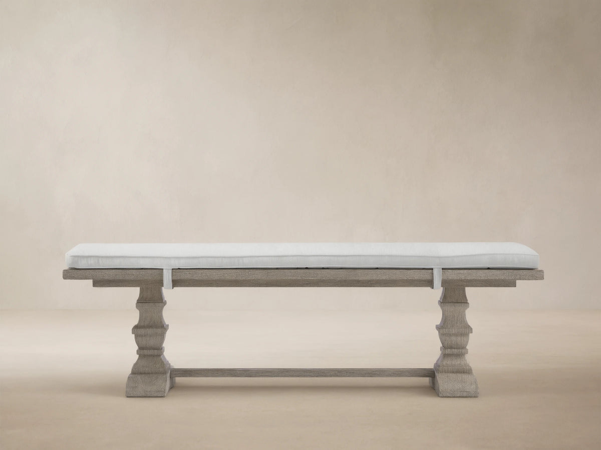 Weathered fawn Hamptons outdoor dining bench, carved pedestals, with a strapped light grey cushion, against a wooden shingle wall.