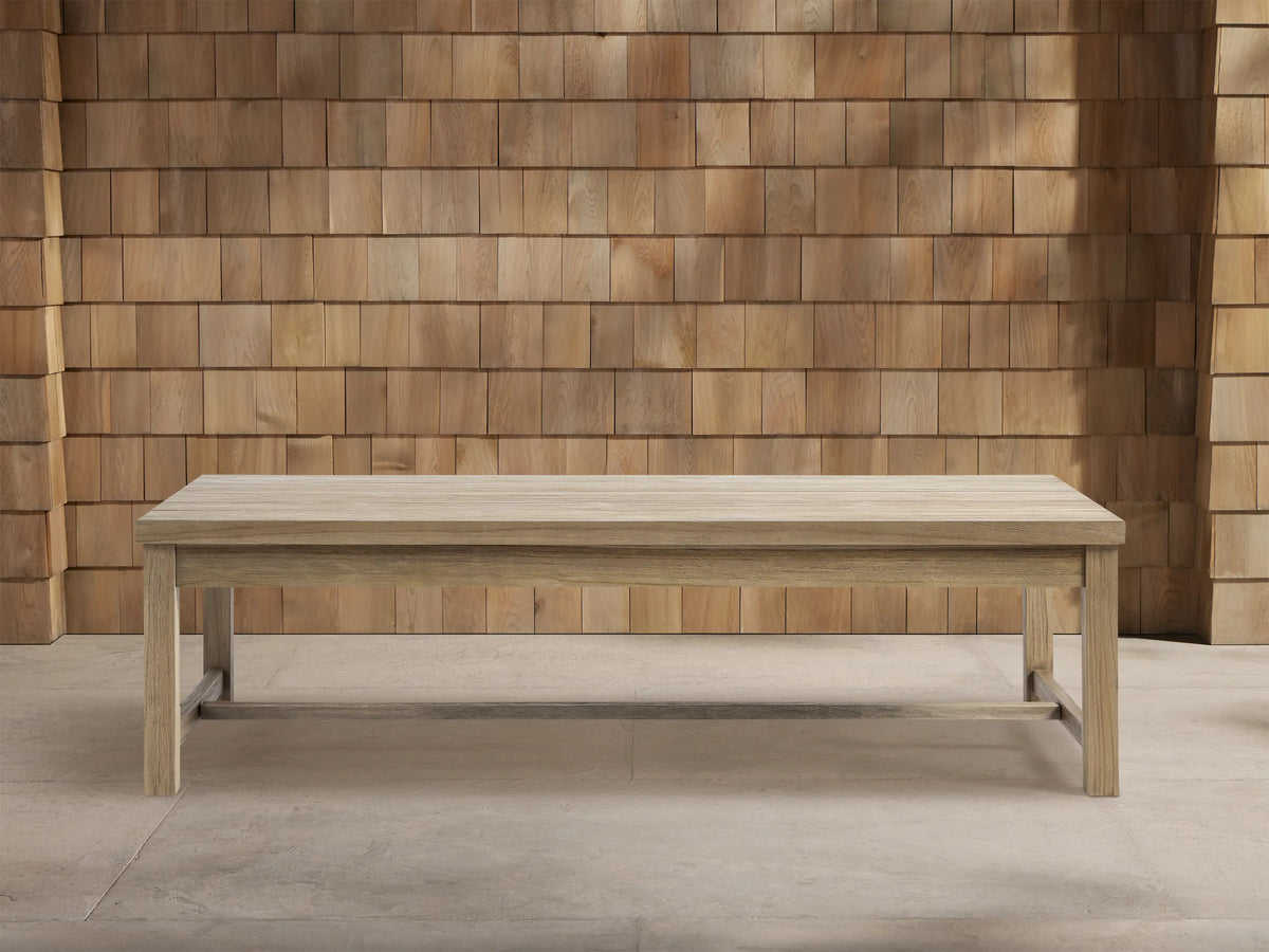 Front view of a rectangular Hamptons Outdoor Coffee Table made of light weathered teak, centered on a gray patio with a wood shingle wall.