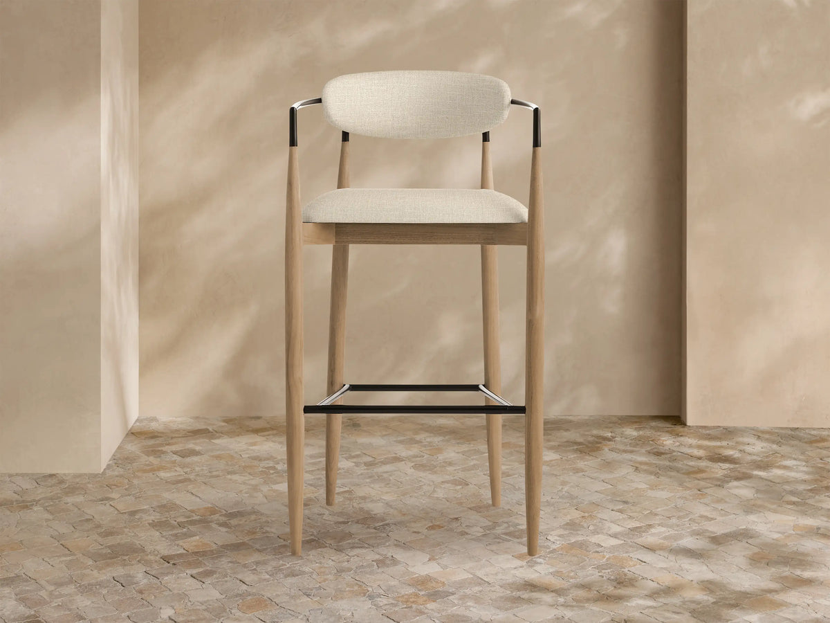 Jagger Outdoor Barstool with light teak wood frame, beige cushioned seat & back, and black metal accents, on a tiled floor.
