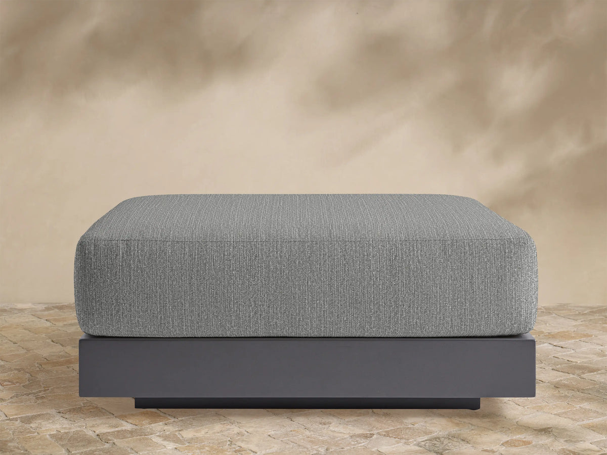 Naxos Outdoor Ottoman, rectangular grey upholstered cushion on a dark grey base, centered on a stone patio with light background.