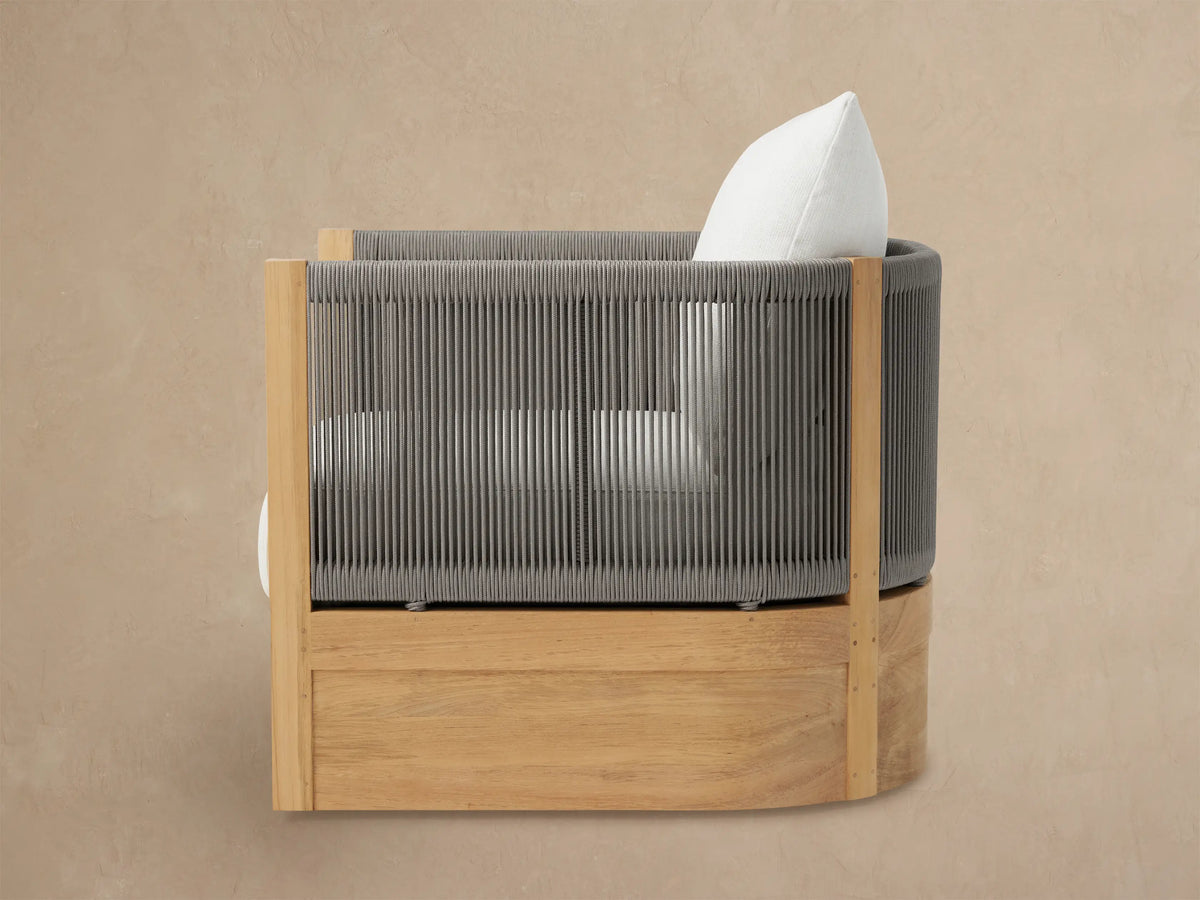 Newport Outdoor Swivel Chair, angled. Natural teak wood base, dark grey woven rope sides, white back cushion on a light wall.