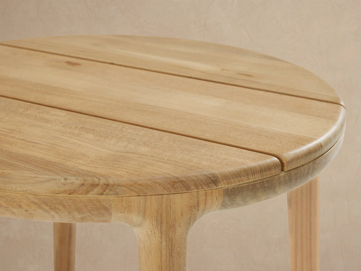 Puglia Outdoor Teak End Table: Round, light natural teak wood table with 4 legs on a tiled patio against a beige wall with shadows.