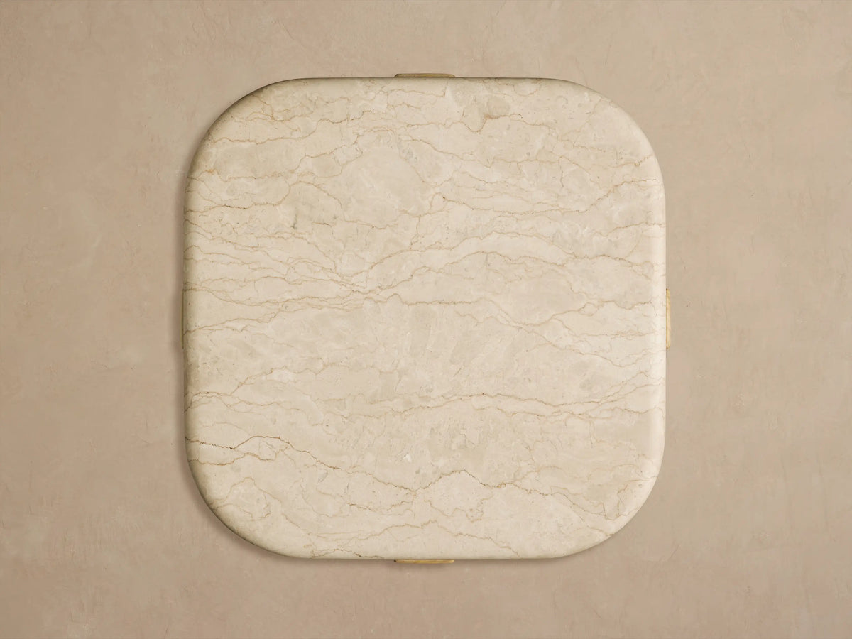 Reily Outdoor End Table. Close-up of its cream marble top with brown veining and rounded edge resting on a light teak wood base.
