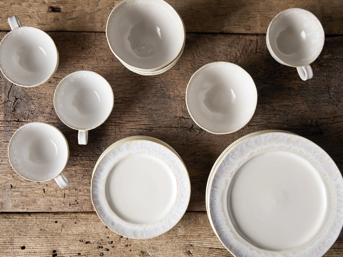 Bela Dinnerware set: Stacked creamy off-white, glazed stoneware bowls & plates, with mugs & saucers on a wooden table.