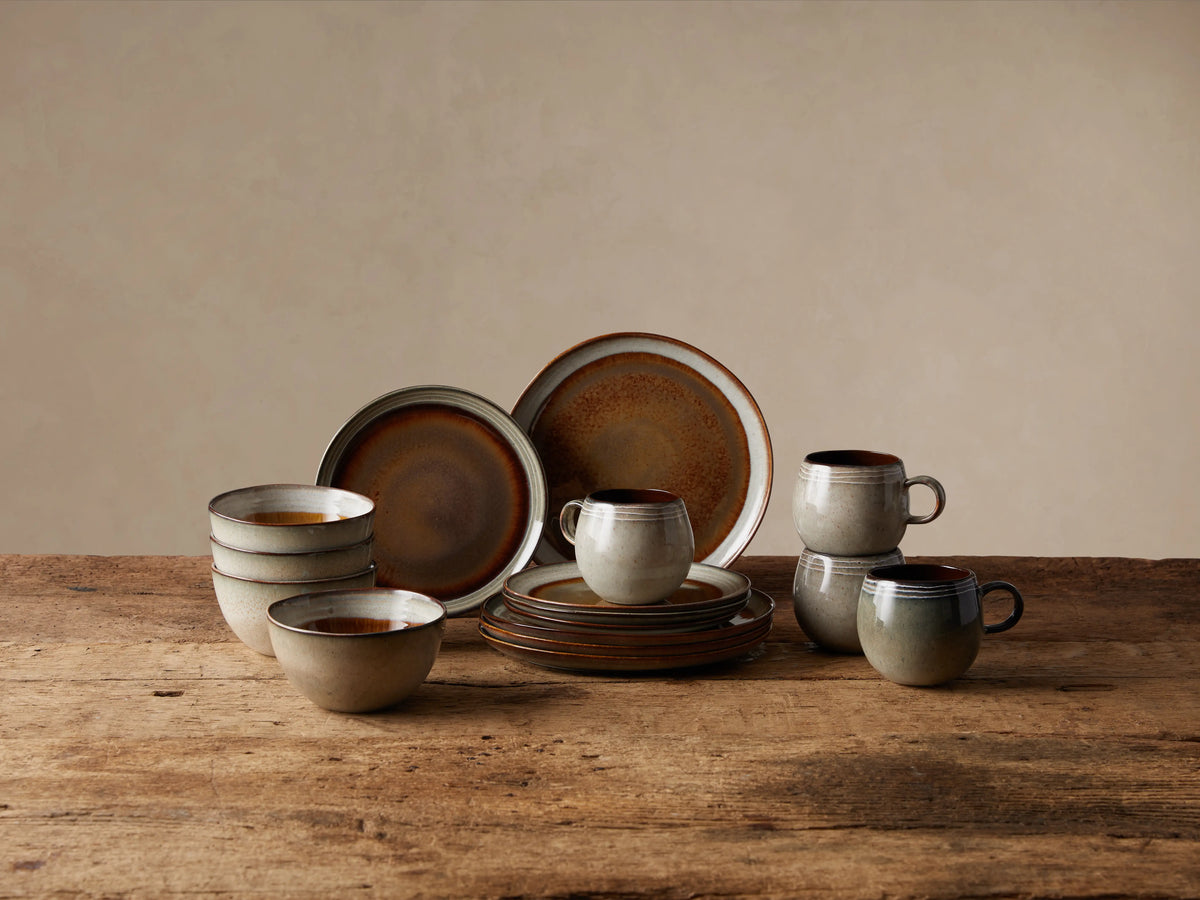Canyon 16-piece dinnerware set on wood table. Reactive hand-glazed stoneware: mugs, bowls, plates in brown & neutral tones.