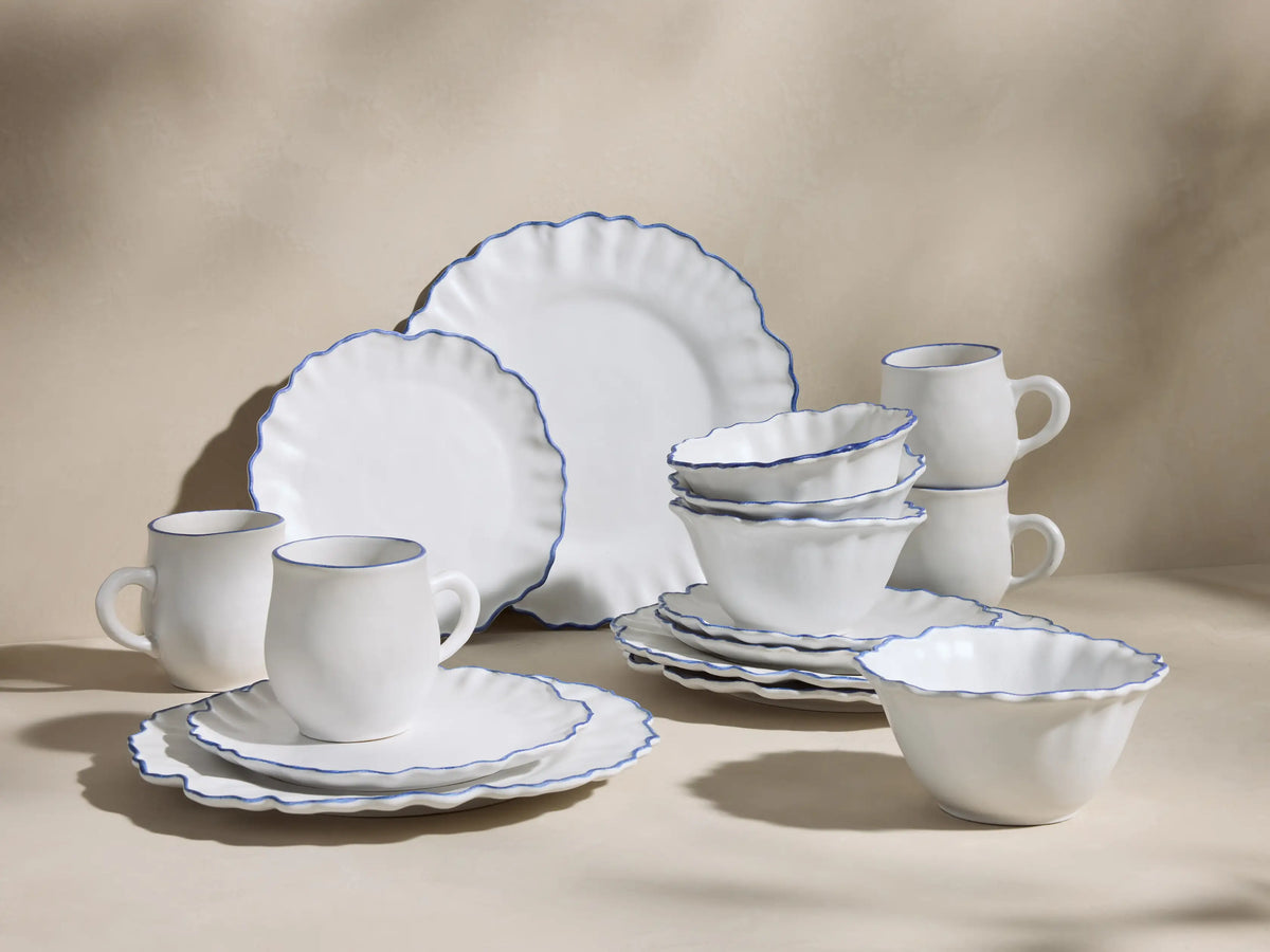 Capuchos dinnerware: White ceramic mug with blue rim, in front of ruffled plates with hand-painted blue edges, angled left.