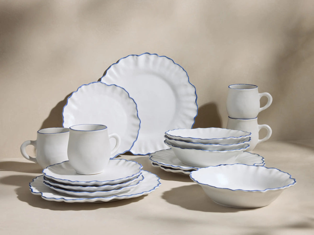 Close-up of white Capuchos ceramic mug with blue rim, next to wavy white plates with blue hand-painted edges.