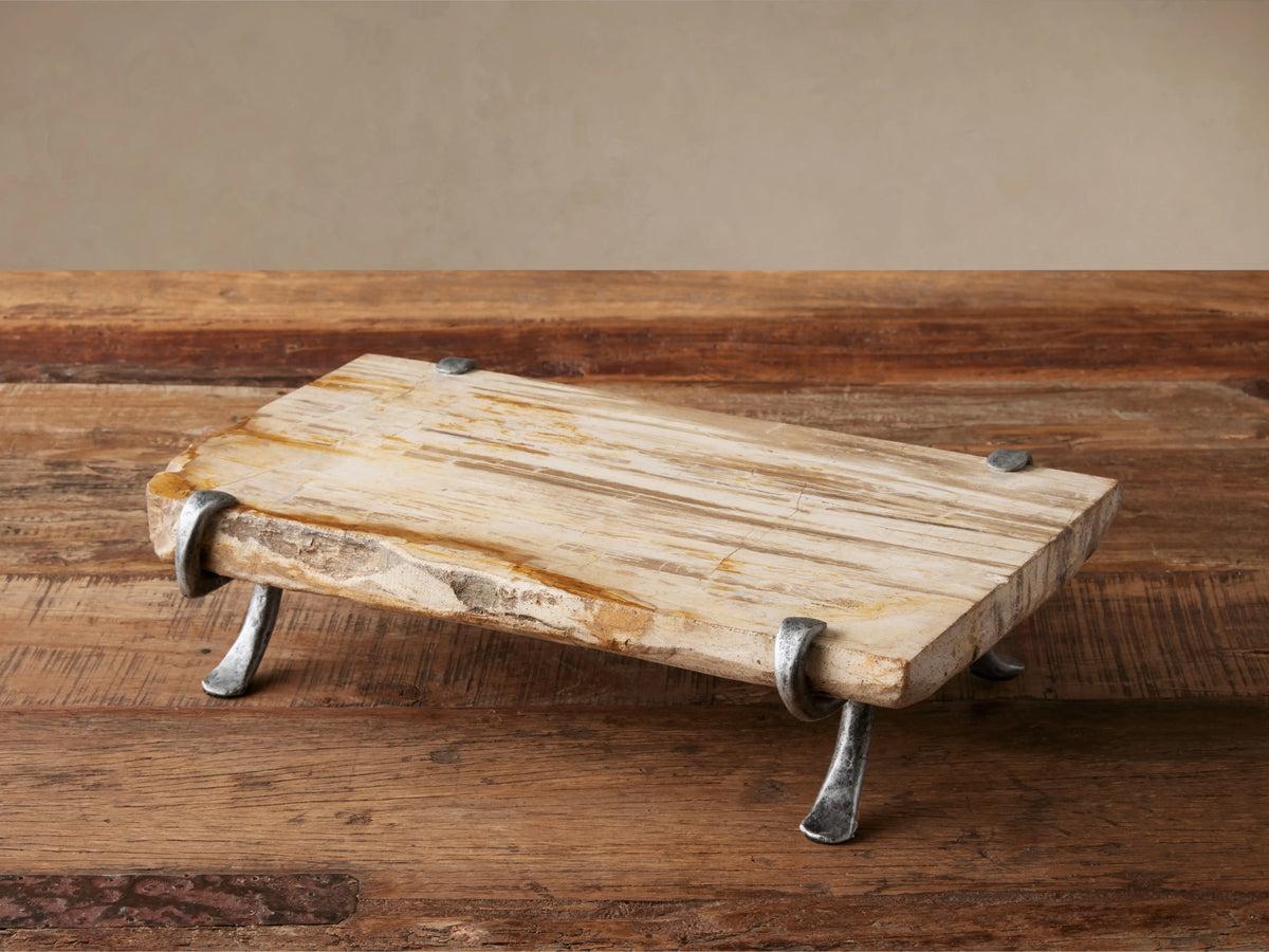 Petrified Wood Serving Board, light-toned with organic edges, held by dark gray wrought iron legs, sitting on a rustic wooden table.
