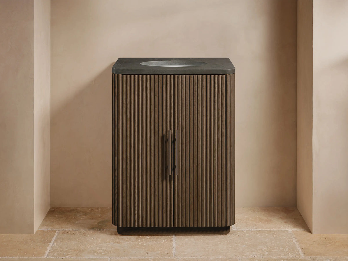 Finnley Powder Vanity's fluted dark wood door slightly open, revealing an antique bronze handle, folded towels & toiletries inside.