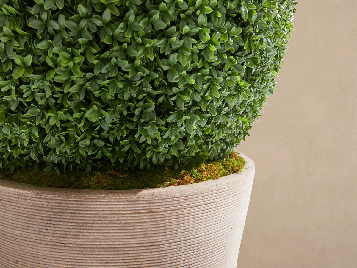 Spherical green faux boxwood in a light beige, ribbed, conical planter, on a stone floor against a tan wall.