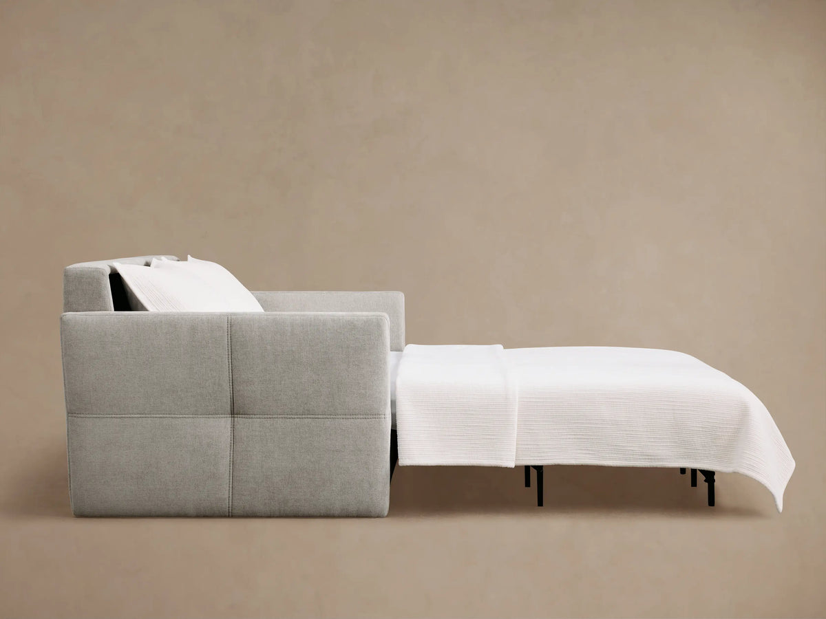 Reece Sleeper Sofa in light gray fabric, partially open to reveal a white mattress and extending upholstered footrest with black legs.