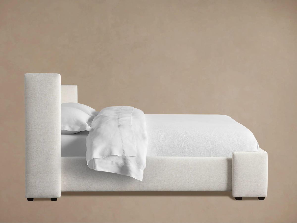 Wyller Bed headboard detail: light off-white textured fabric, showing a clean, padded corner against a warm beige wall.