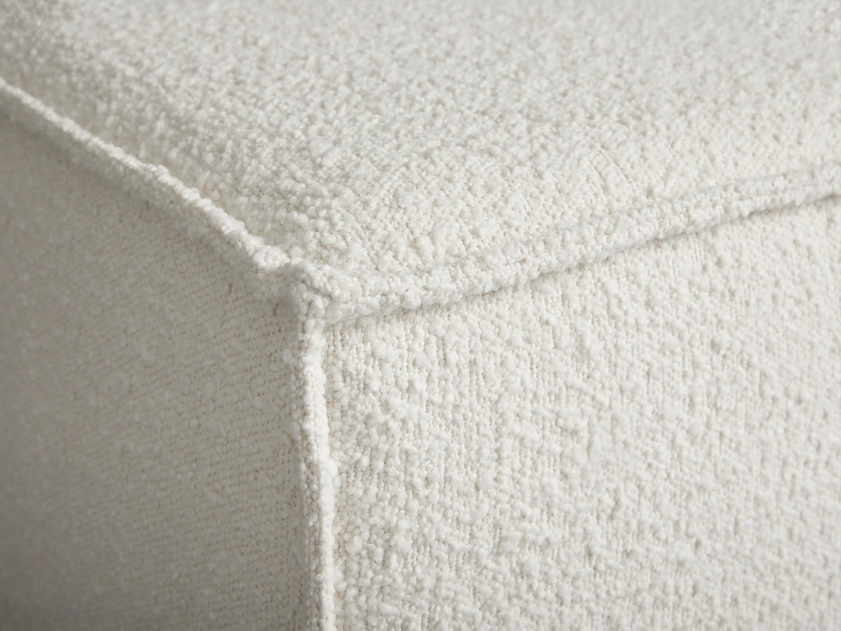 Rectangular Gabe Ottoman in cream bouclv© fabric with piping, on a wooden floor against a light beige wall.