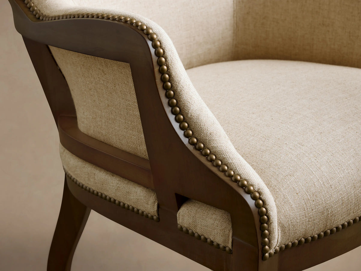 Front 3/4 view of the light beige upholstered Tilly Chair, featuring a dark brown wooden frame and brass nailhead detailing.