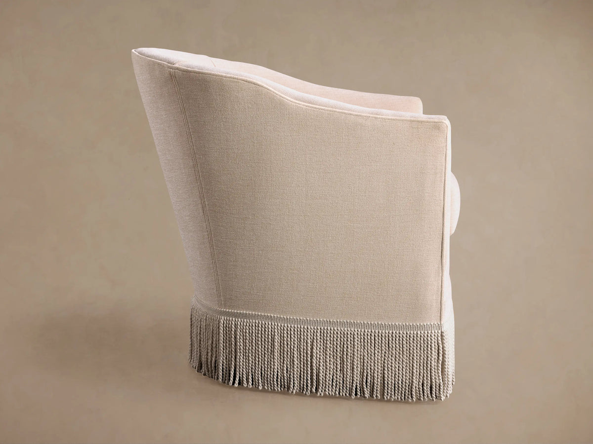 Back-side view of a light beige Giles Swivel Chair. Textured upholstery, vertical seam, and thick, tasseled fringe along the bottom.