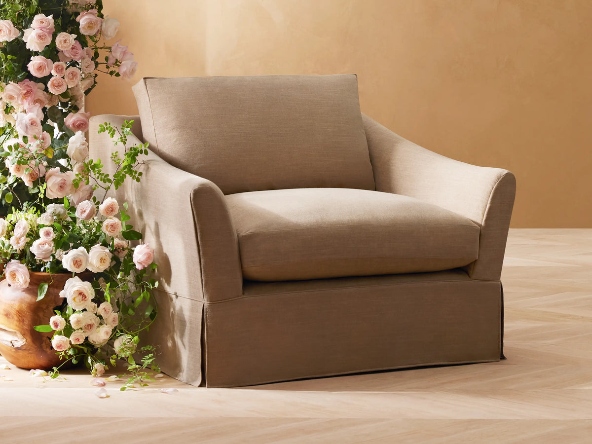 Tan Helena Slipcovered Swivel Chair with soft cushions and a tailored skirt, placed beside a pot of pink and white flowers on wood floor.