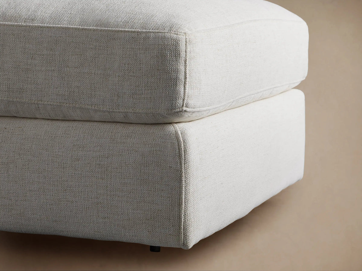 Beale Ottoman, a plush, light-toned upholstered ottoman with a soft, rounded cushion on a solid base, on a wood floor.