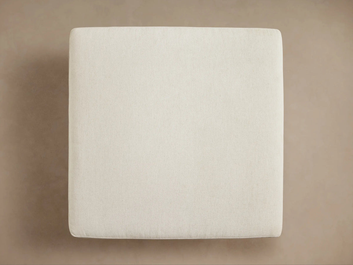 Interior view of an open cream upholstered storage ottoman, revealing a folded brown and cream plaid throw blanket with fringe inside.