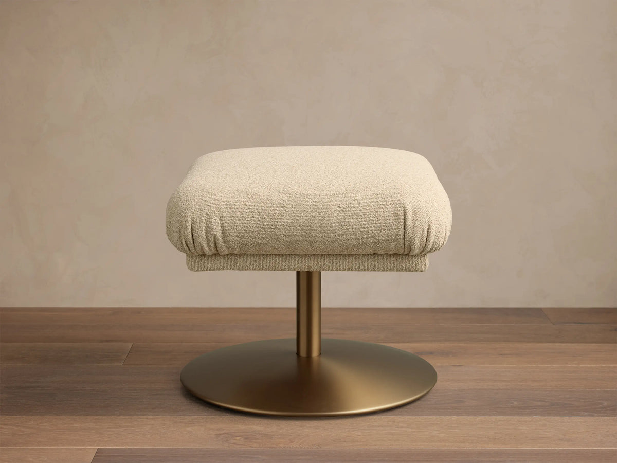 Front view of a square beige fabric upholstered ottoman with a golden pedestal base on a wooden floor.