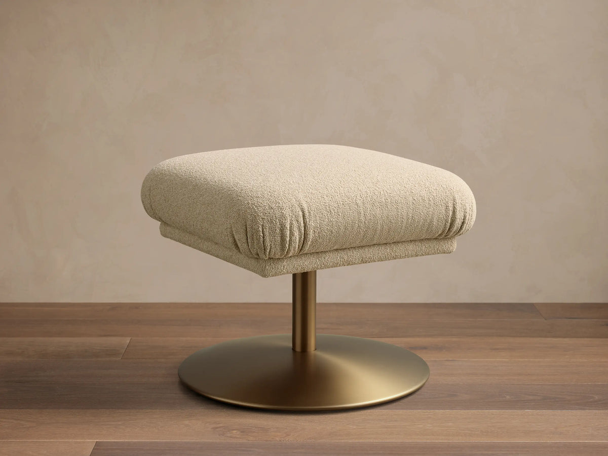 Front view of a square beige fabric upholstered ottoman with a golden pedestal base on a wooden floor.
