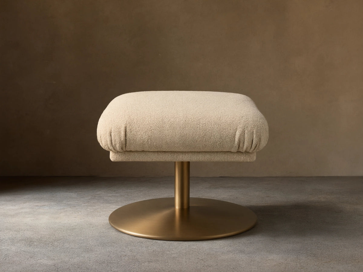 Front view of a square beige fabric upholstered ottoman with a golden pedestal base on a wooden floor.