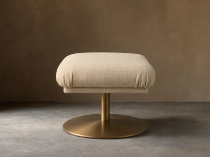 Front view of a square beige fabric upholstered ottoman with a golden pedestal base on a wooden floor.