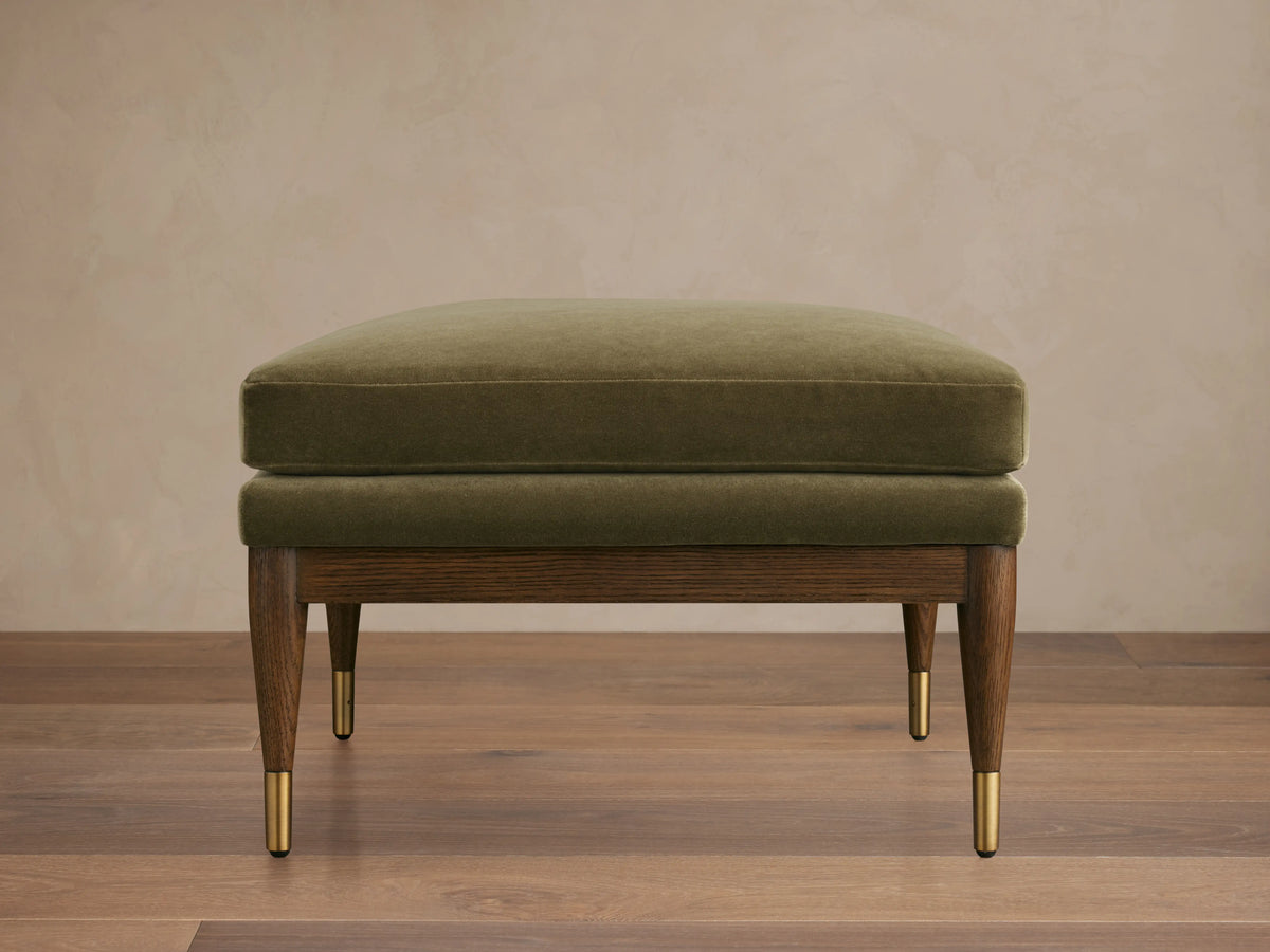 Olive green Fisher Square Ottoman with a plush cushion, mocha-finished red oak legs, and brass caps, on a wooden floor.