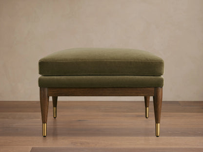 Olive green Fisher Square Ottoman with a plush cushion, mocha-finished red oak legs, and brass caps, on a wooden floor.