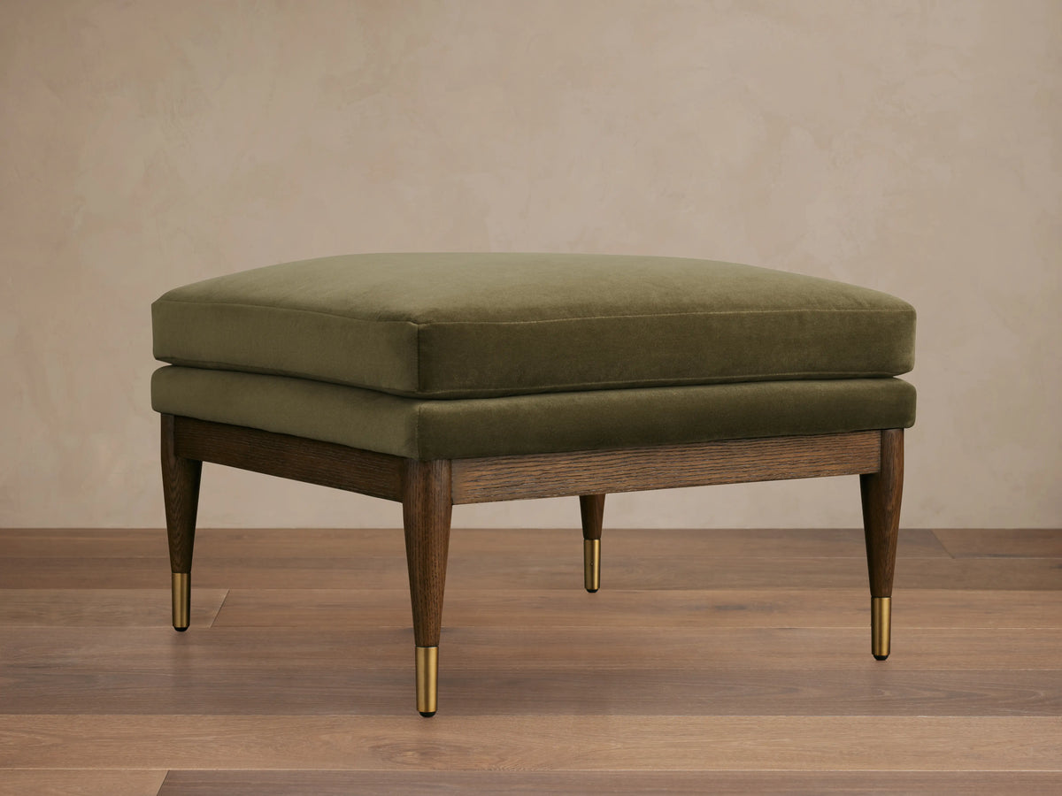 Olive green Fisher Square Ottoman with a plush cushion, mocha-finished red oak legs, and brass caps, on a wooden floor.