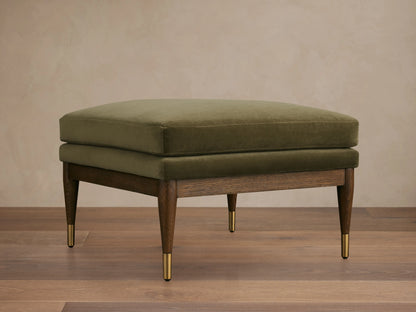 Olive green Fisher Square Ottoman with a plush cushion, mocha-finished red oak legs, and brass caps, on a wooden floor.
