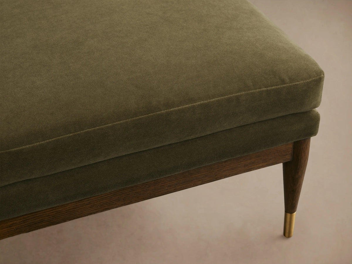 Square ottoman with green cushioned top, wooden frame in mocha finish, brass-tipped tapered legs on wood floor.
