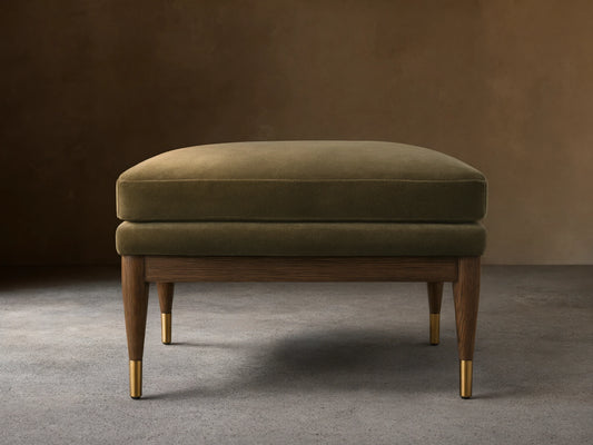 Olive green Fisher Square Ottoman with a plush cushion, mocha-finished red oak legs, and brass caps, on a wooden floor.