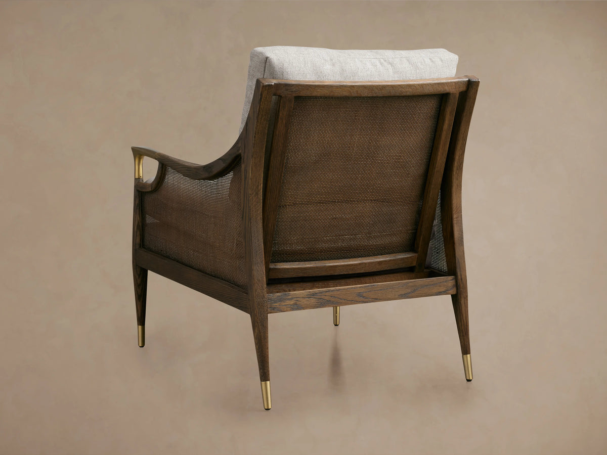 Cane back chair with mocha-finished oak frame, gold metal arm accents, gray cushioned seat, and textured fabric upholstery.